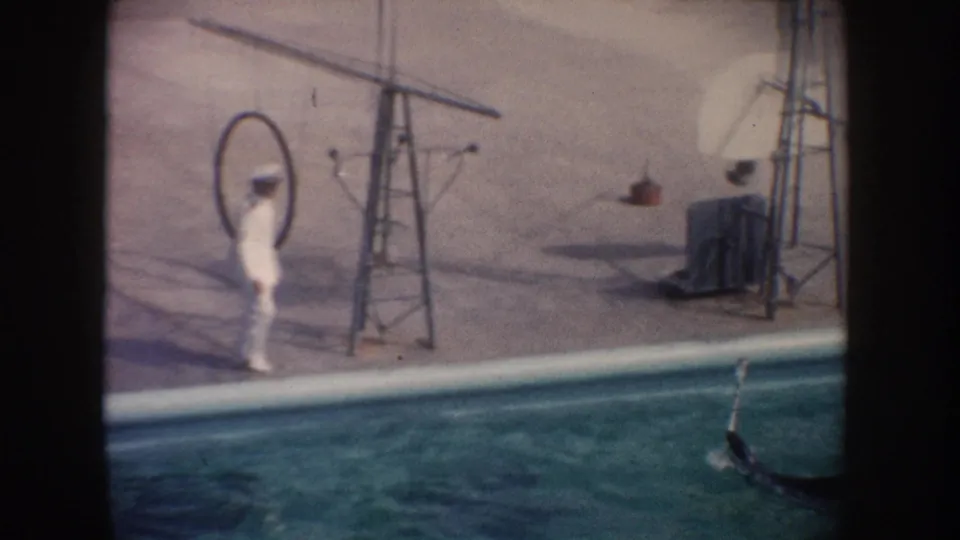 1962 California home-movie frame showing a white-uniformed trainer at poolside with a hoop, poles, and a seal partly visible in the water.