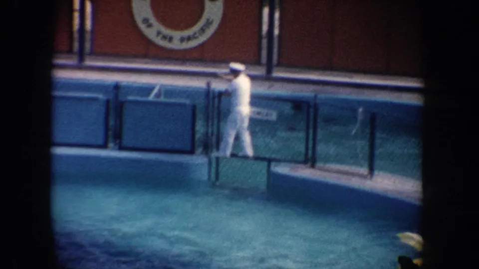1962 California aquarium frame with a trainer in white walking along the pool edge beneath partial signage reading "OF THE PACIFIC."