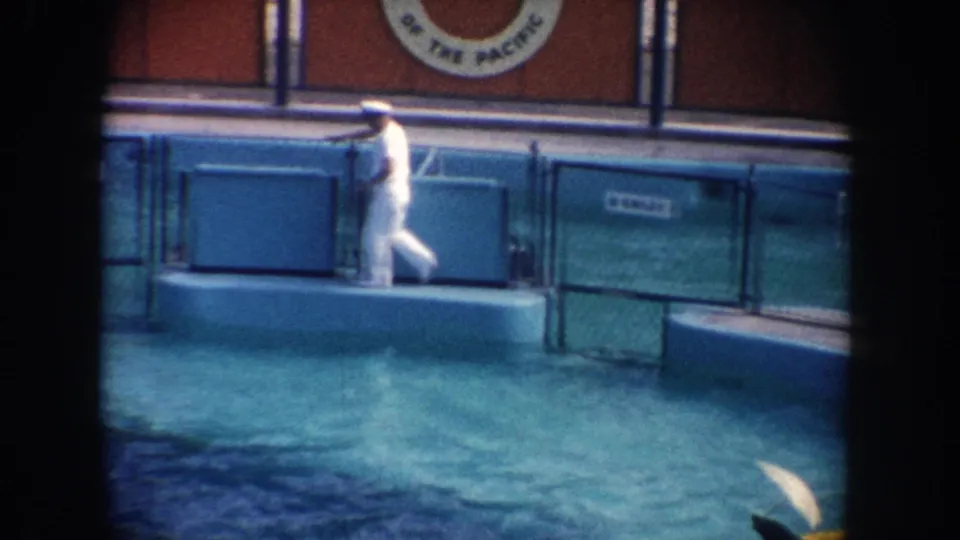 1962 California home-movie frame showing the trainer paused on a low platform beside the aquarium pool and blue show barriers.