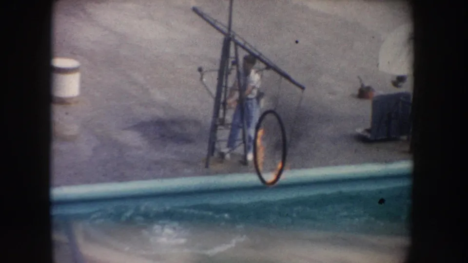 1962 California home-movie frame with the handler at the metal rig and flaming hoop beside the dolphin pool.