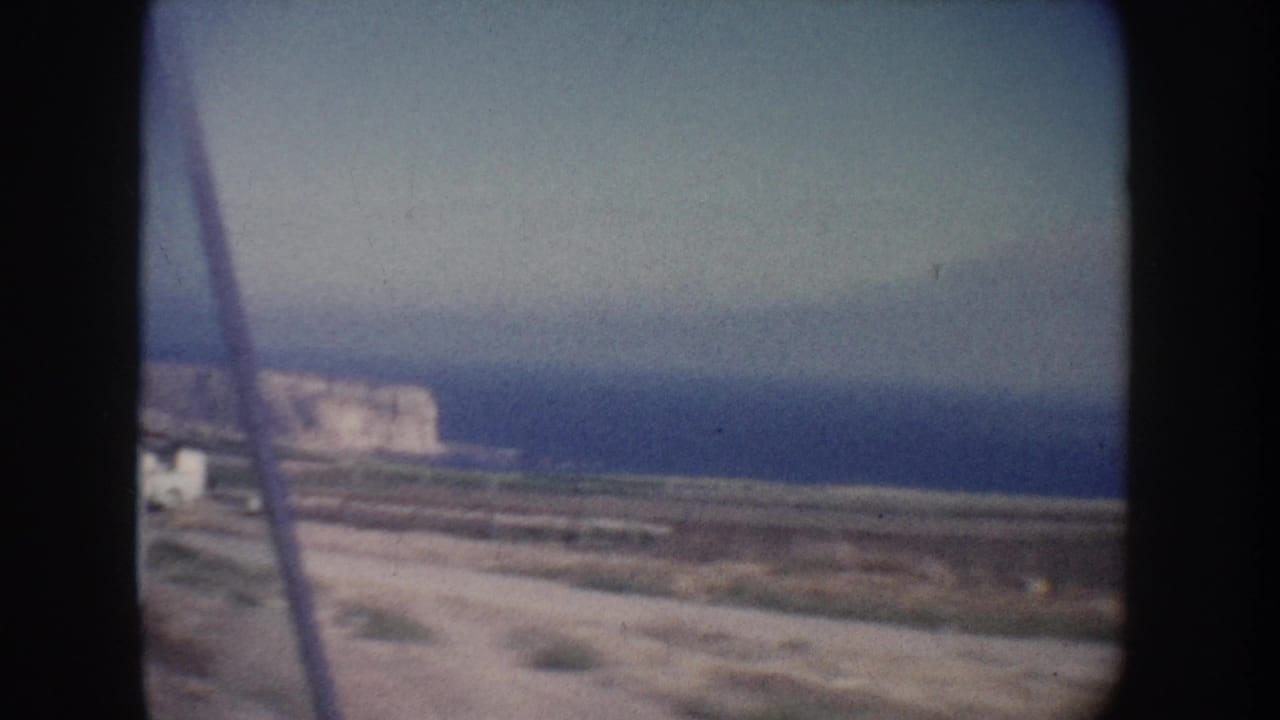 1962 California coastal view from a moving vehicle, with a pale cliff face, blue ocean, dirt roadside, and film gate shadows.