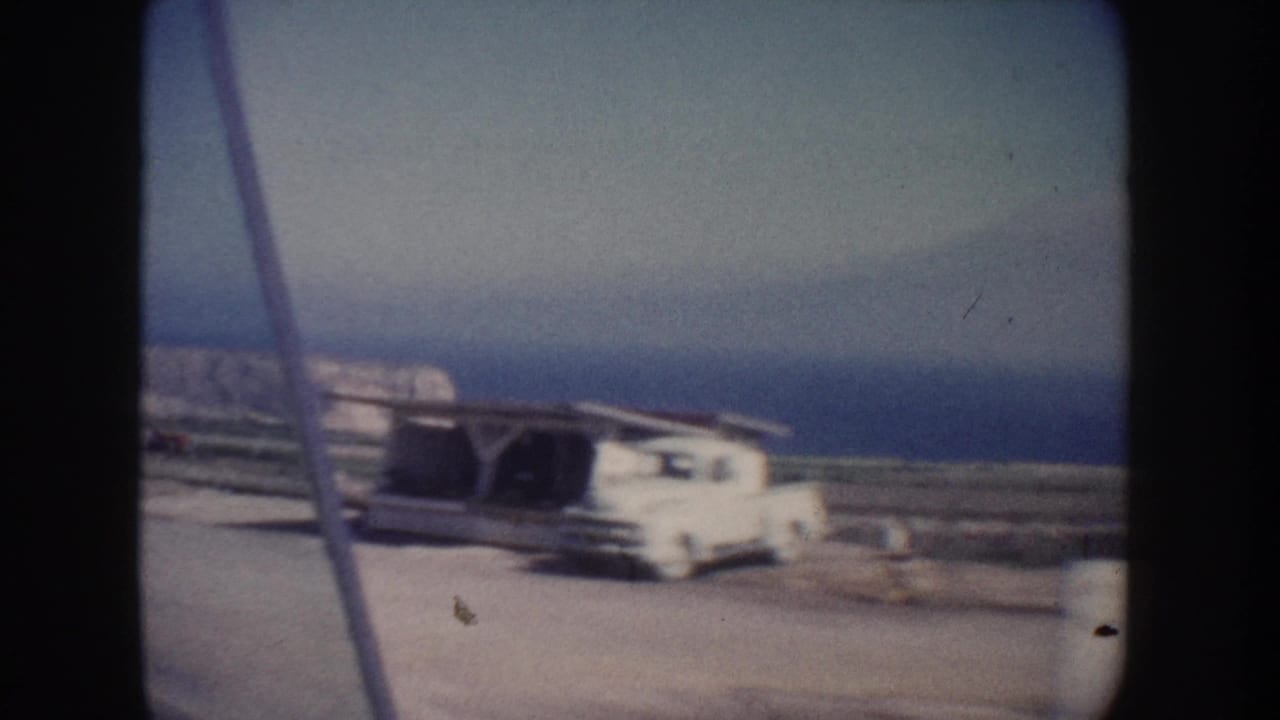 A white vintage truck appears parked near a low shed along a California coastal road, with ocean and cliff landscape behind it.