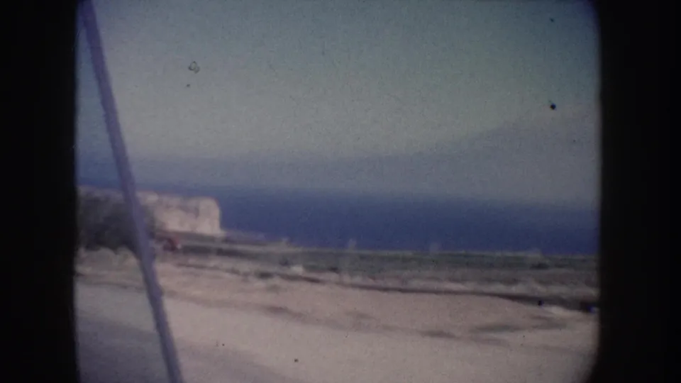 The moving camera looks past a roadside edge toward the California coast, showing a pale cliff, blue water, and soft 8mm blur.