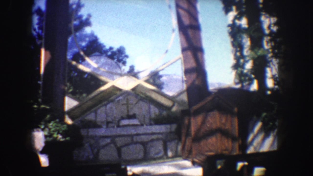 1962 California home-movie frame with a large archway, stone religious feature, cross, trees, and visible dark film borders.