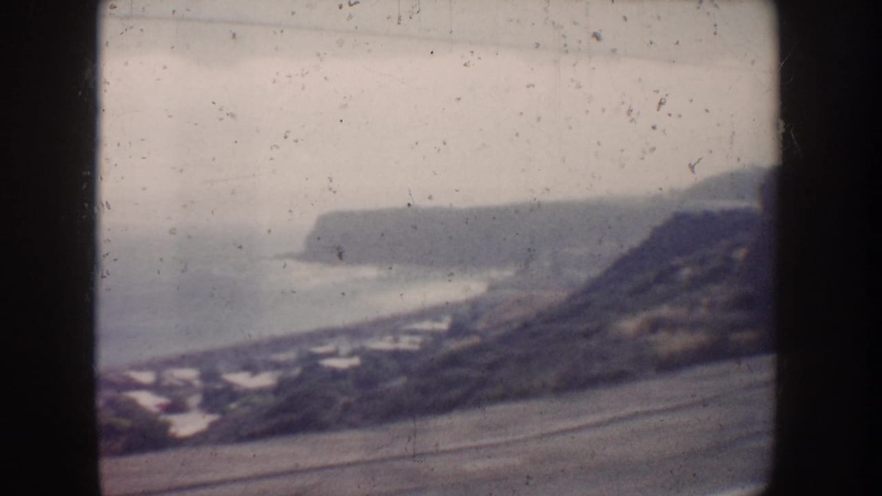 Hazy coastal view with ocean, beach, bluffs, houses, and a road seen through dark film borders.