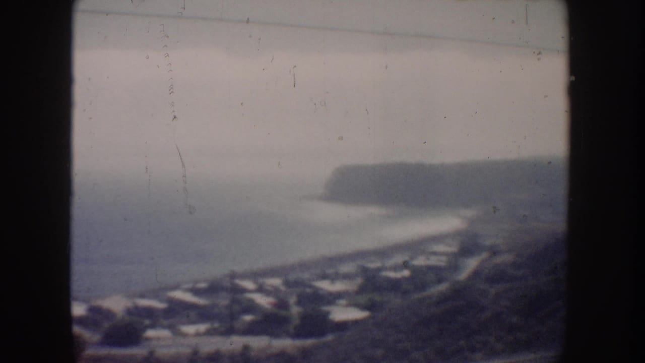 1962 California coastal home-movie frame with a bay-like shoreline, distant headland, beach curve, houses, and soft overcast haze.