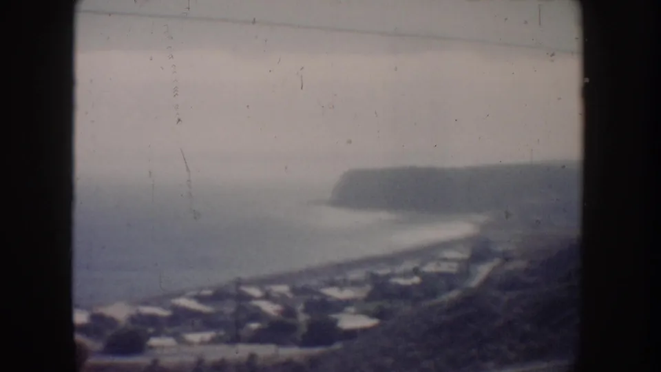 1962 California coastal home-movie frame with a bay-like shoreline, distant headland, beach curve, houses, and soft overcast haze.