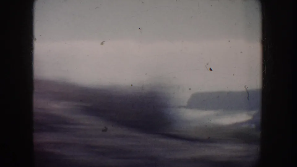 Motion-blurred 1962 California coast frame showing water, beach, distant headland, and dark foreground obstruction from a moving viewpoint.