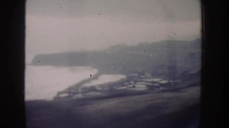1962 California 8mm frame showing a hazy elevated view of beach, ocean water, houses, hillside, and distant coastal landform.
