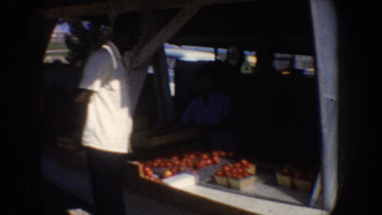 Man at a roadside fruit stand with crates of red produce beside a vehicle or stall.