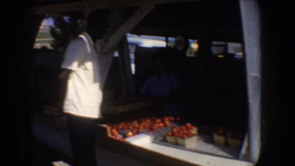 1962 California roadside fruit stand scene with a man in a white shirt and crates of red produce.