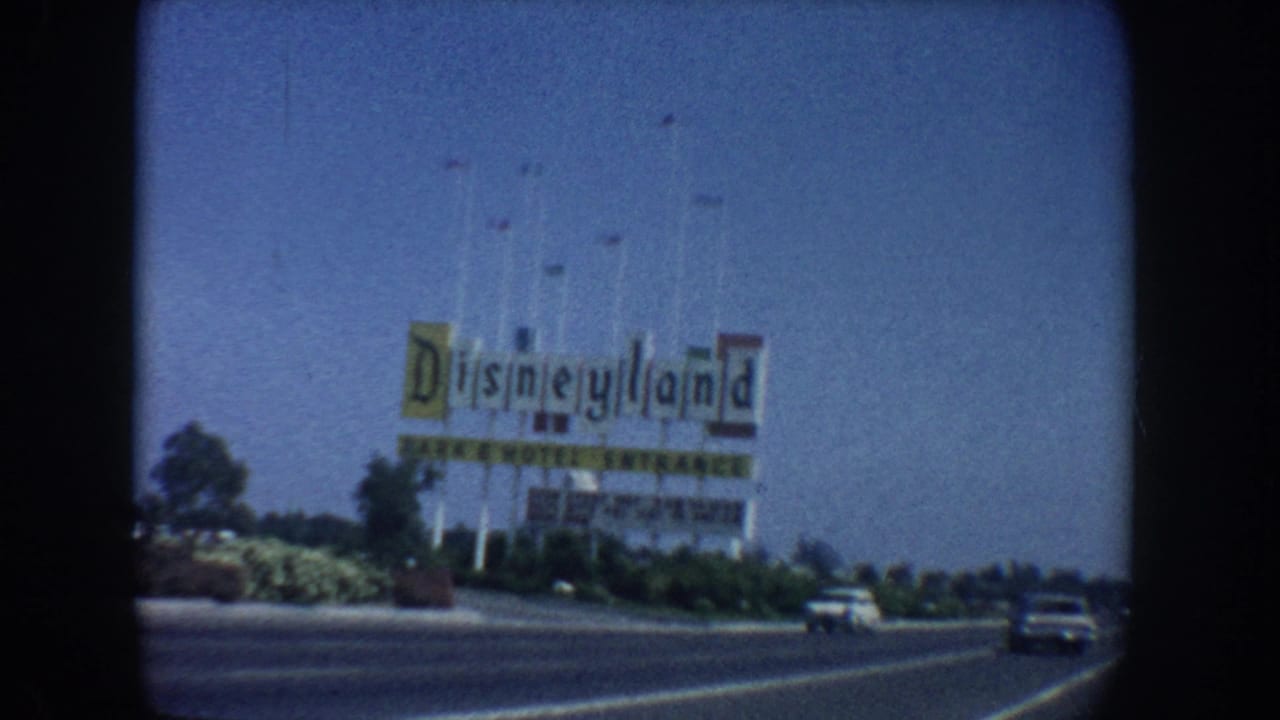 1962 California 8mm frame showing the Disneyland Park and Hotel Entrance sign in the distance from a moving car, with roadway, landscaping, and early-1960s vehicles visible.
