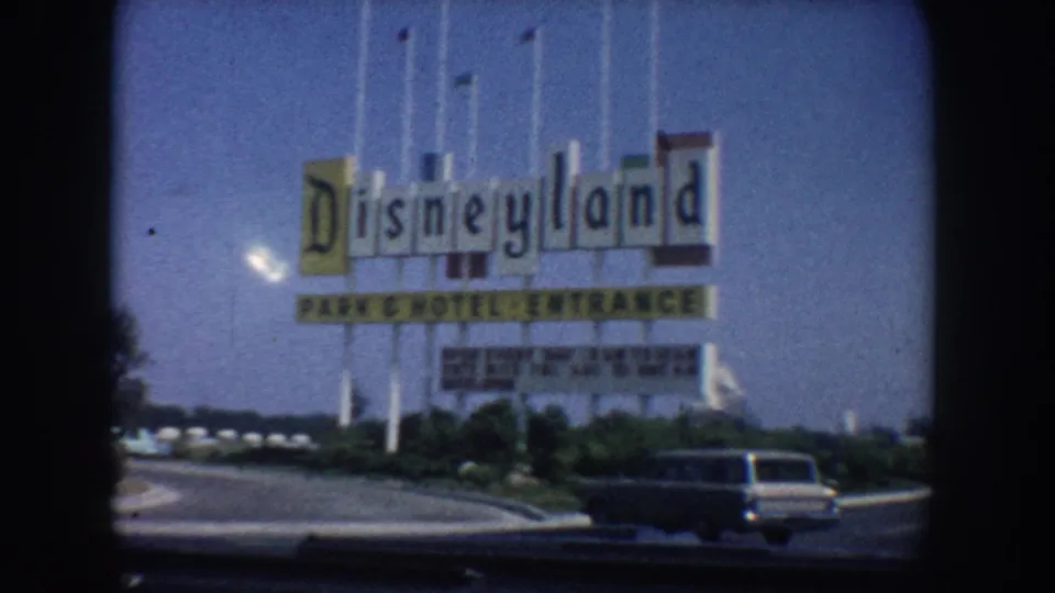 Closer 1962 California home-movie view of the Disneyland Park and Hotel Entrance sign, roadside landscaping, flagpoles, and a dark station wagon or sedan near the foreground.