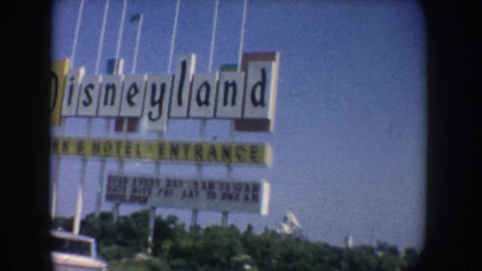 Tight 1962 California frame of the Disneyland sign filling much of the view, with Park and Hotel Entrance lettering, flagpoles, shrubs, and blue sky visible through soft 8mm motion blur.