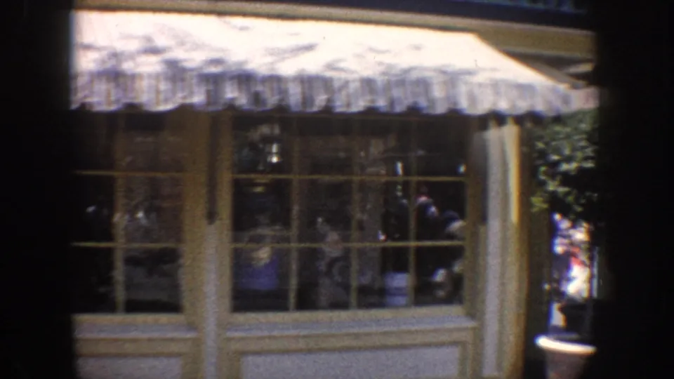 Storefront window under a striped awning in Anaheim, with reflected figures and passing visitors in soft 8mm focus.