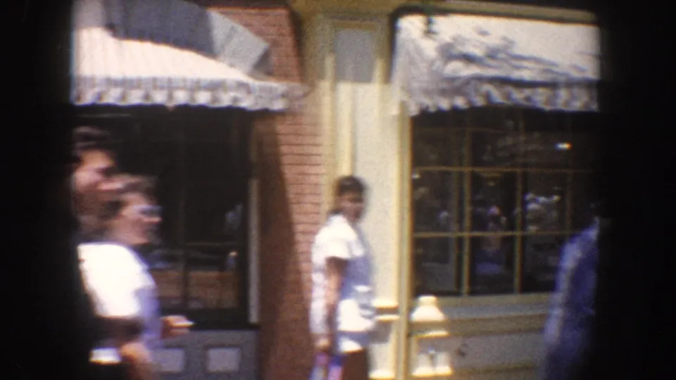 People walk past Anaheim storefront windows and shaded awnings, captured from a handheld 1962 8mm perspective.