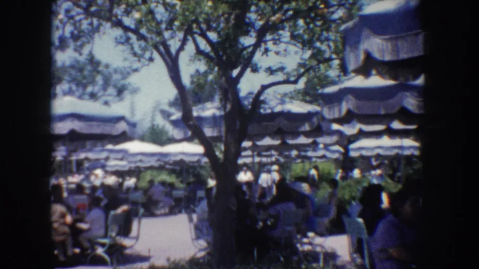 1962 Anaheim outdoor dining area with visitors seated beneath striped umbrellas and trees in soft 8mm color.
