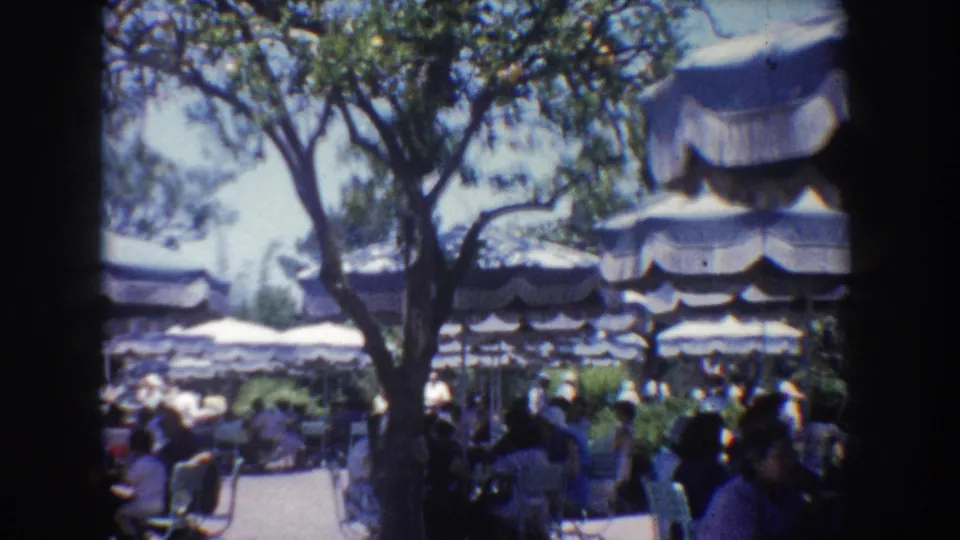 Crowded Anaheim patio scene with trees, striped umbrellas, and visitors seated or standing in a shaded leisure area.