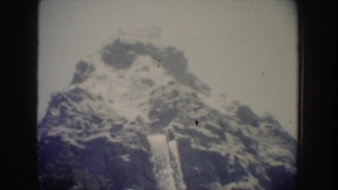 1962 Anaheim 8mm home-movie frame showing a pale waterfall dropping down a rugged, snow-edged rocky mountain facade.
