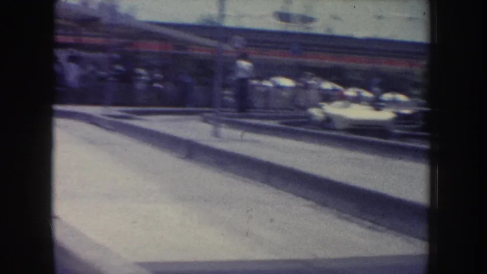 1960s Anaheim home-movie frame with an elevated rail structure, red-and-blue vehicle, walkway, and distant parked cars.