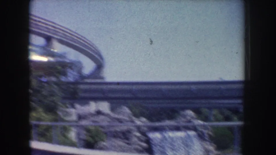 Visitor-shot 8mm frame from Anaheim showing a curving elevated structure over landscaped rocks, greenery, and water.
