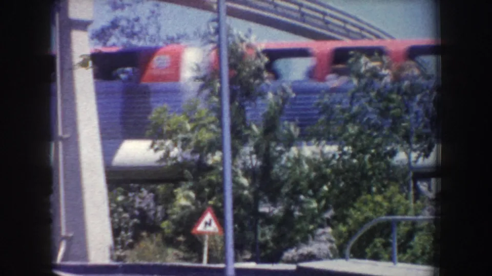 1960s Anaheim home-movie frame with a red-and-blue elevated transit vehicle behind trees, poles, railings, and landscaped planting.