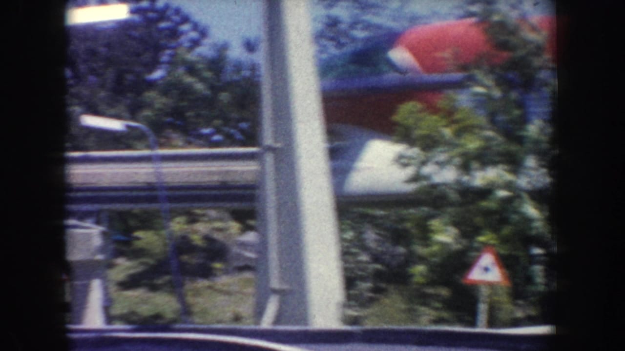 1962 Anaheim 8mm frame showing a red and blue monorail partly obscured by trees, poles, and a triangular roadside warning sign.