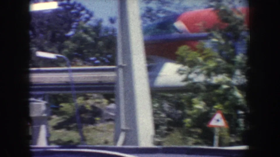 1962 Anaheim 8mm frame showing a red and blue monorail partly obscured by trees, poles, and a triangular roadside warning sign.