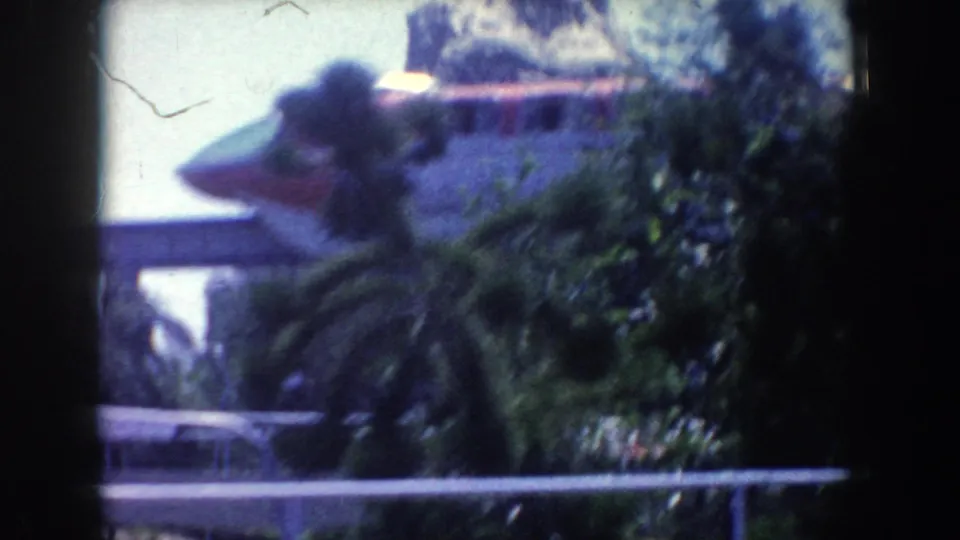 1962 Anaheim home-movie frame with an elevated track, dense roadside greenery, railing, and motion blur from a passing vehicle.