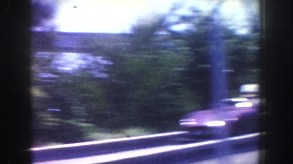 1962 Anaheim 8mm frame of a blurred roadside scene with guardrail, greenery, bright sky, and a passing car shape near the frame edge.