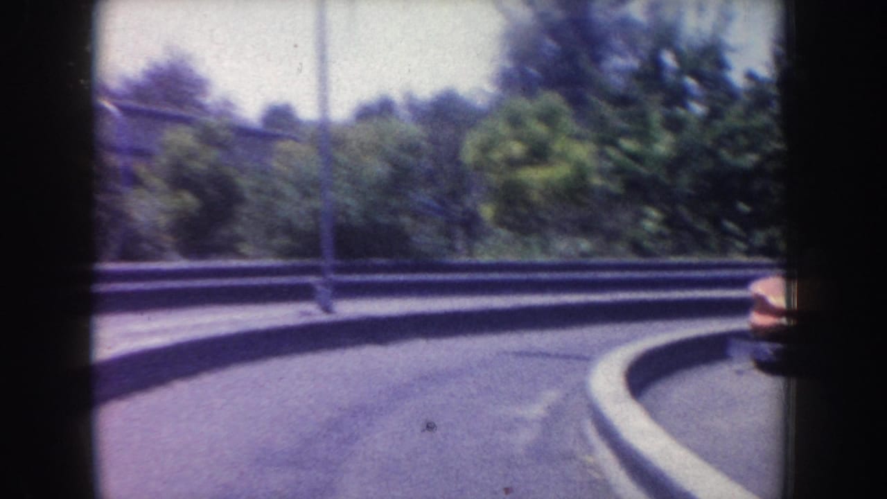 1962 Anaheim home-movie frame showing a curved park roadway, roadside planting, trees, and a partial classic car passing at frame edge.