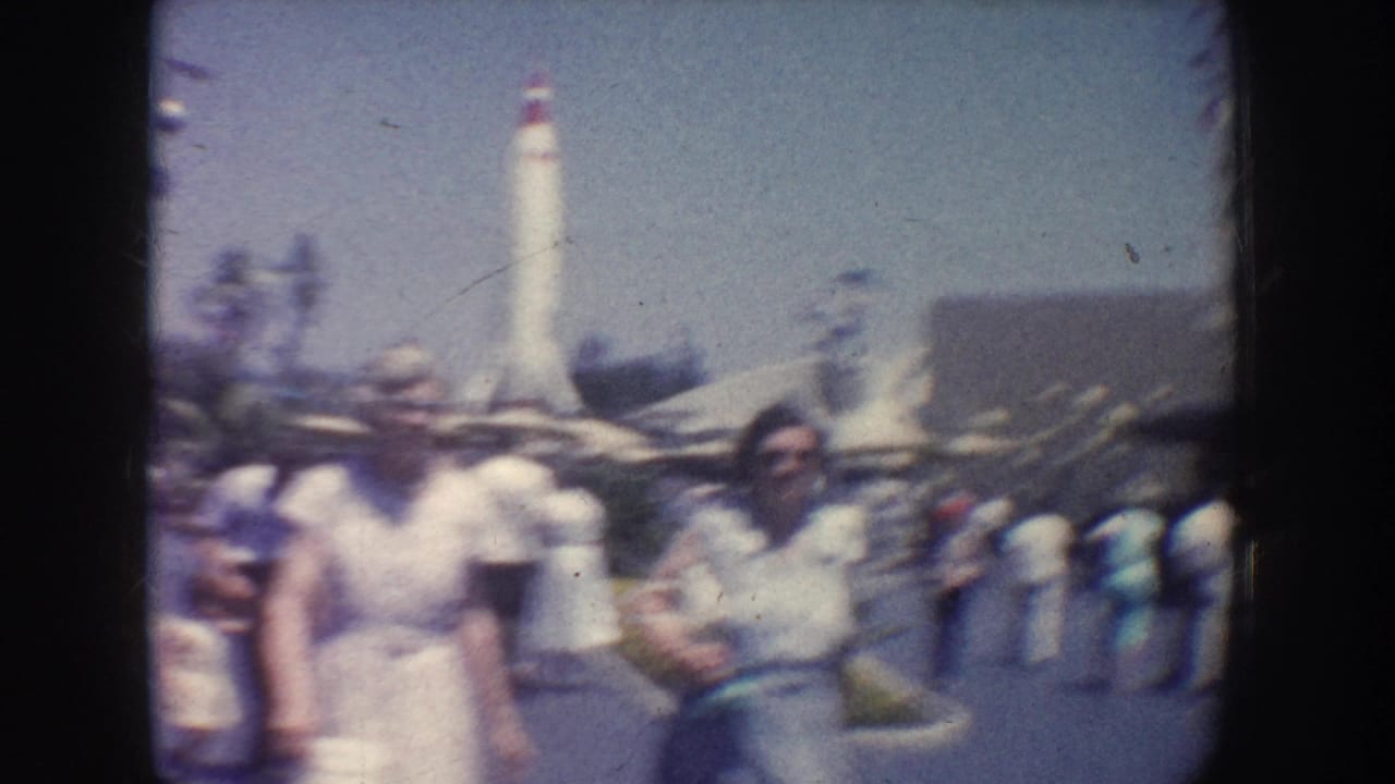 Visitors stand near a tall white rocket exhibit in a sunny amusement park.