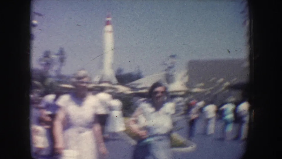 1962 Anaheim frame with mid-century visitors in front of a tall white rocket exhibit, palm trees, and park structures under blue sky.