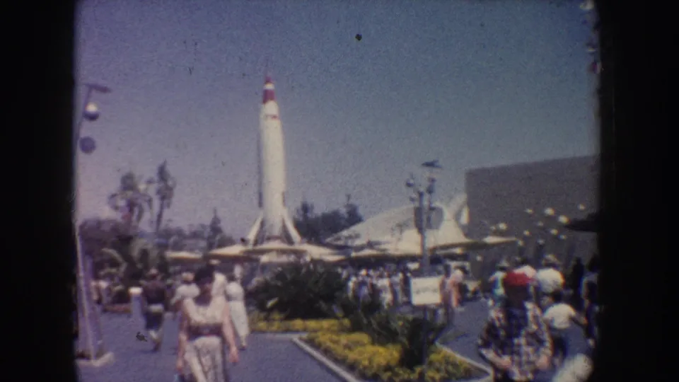 1962 Anaheim home-movie frame showing visitors, flower beds, lampposts, palm trees, and a white rocket exhibit in the distance.