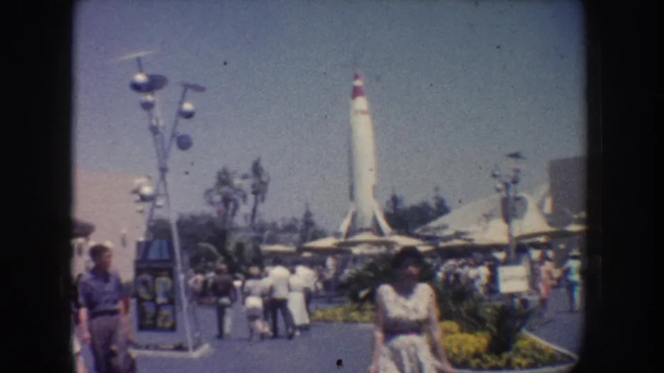 1962 Anaheim frame showing a busy walkway, visitors in period clothing, futuristic lampposts, landscaping, and a tall white rocket display.