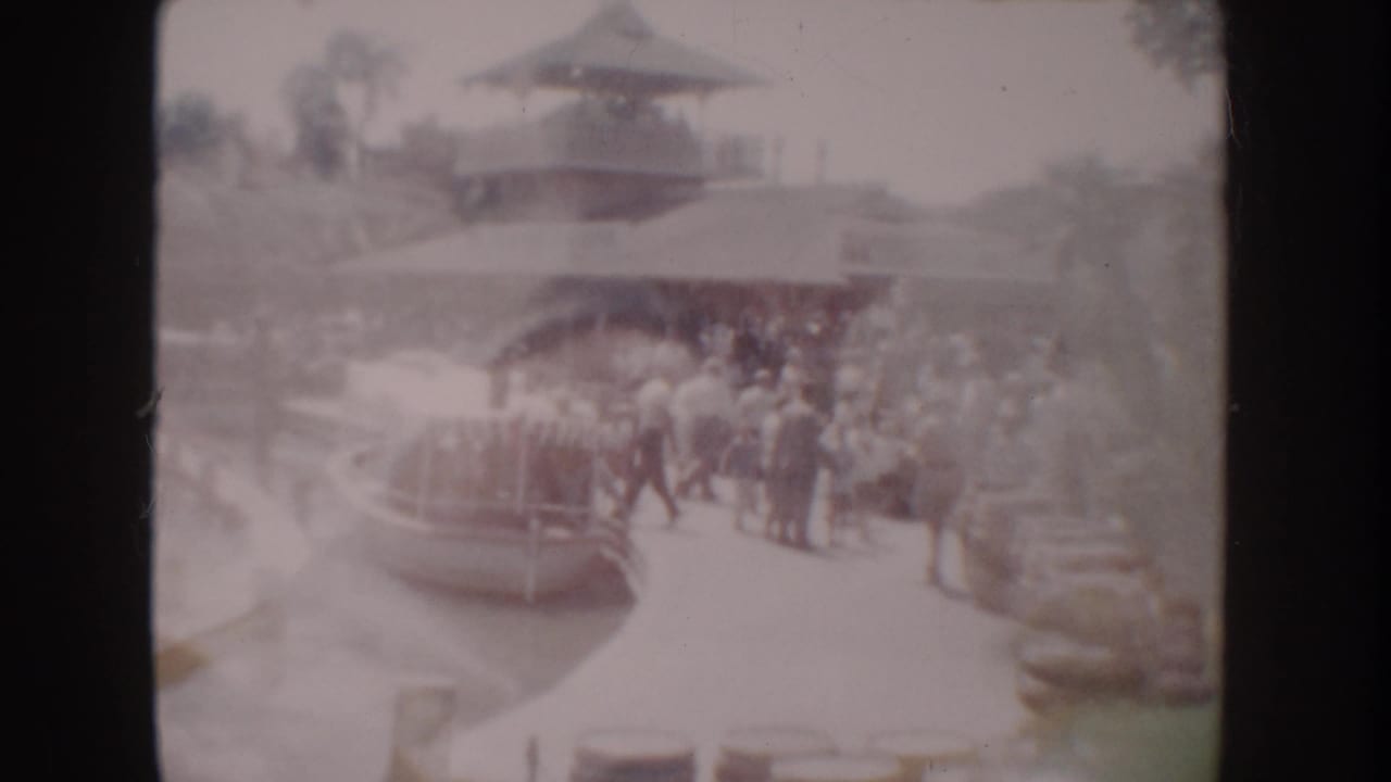 1962 Anaheim home-movie frame showing visitors gathered by a small boat at a landscaped dock with a pagoda-style roof behind them.