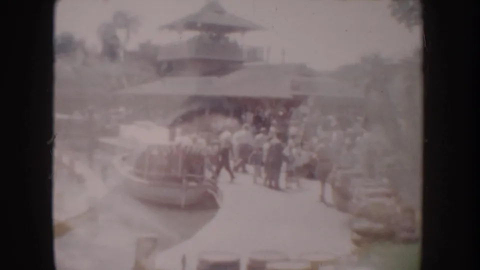 1962 Anaheim home-movie frame showing visitors gathered by a small boat at a landscaped dock with a pagoda-style roof behind them.