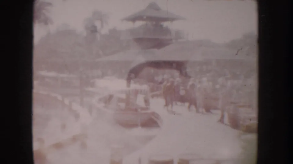 1962 Anaheim home-movie frame showing visitors beside a docked boat, with water, landscaping, and a pagoda-style roof in the background.