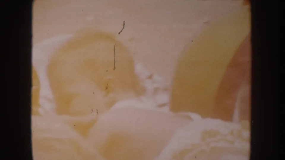 Faded 1962 Anaheim home-movie frame appears to show a baby in light clothing or a sundress beside pale bedding or fabric.