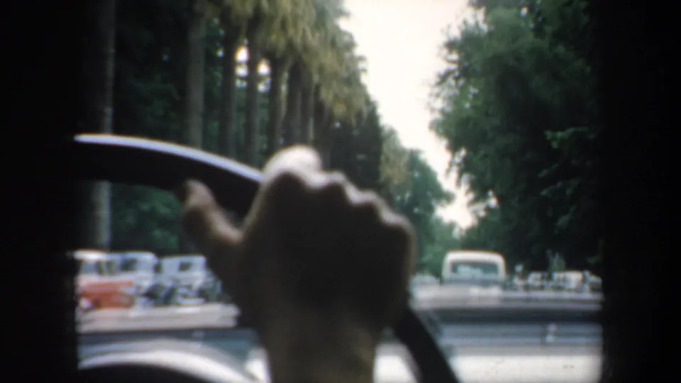 1957 San Francisco 8mm frame from inside a moving car, with a hand on the steering wheel and mid-century cars ahead on a palm-lined street.