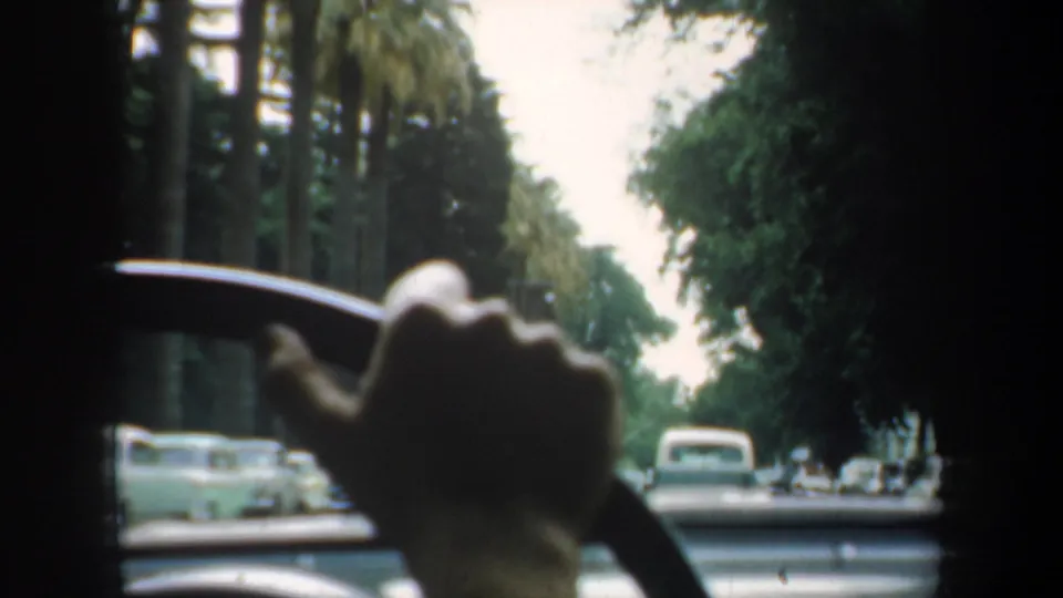 1957 San Francisco in-car 8mm frame showing a silhouetted hand on the steering wheel, classic cars ahead, and tall palms along the road.