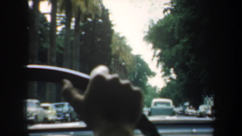 1957 San Francisco home-movie frame looking forward from inside a car toward a tree-lined street with parked mid-century automobiles.