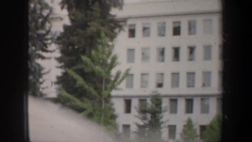 1957 8mm travel frame showing a white civic-looking building partly obscured by trees and a dark vehicle window frame.