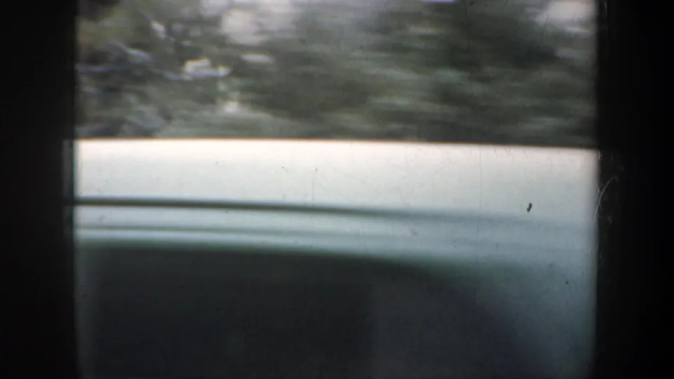 Soft 8mm frame from a moving vehicle, with dark window framing, a pale horizontal surface, and blurred greenery beyond.