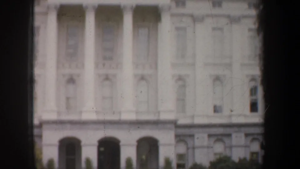 1957 8mm frame showing a white classical civic facade with tall columns, arched lower openings, and trees at the base.
