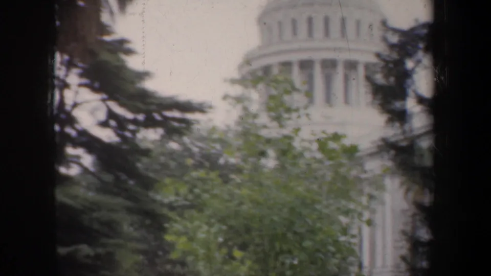 The California State Capitol is partially hidden by green foliage and dark tree silhouettes in a soft 1950s 8mm frame.