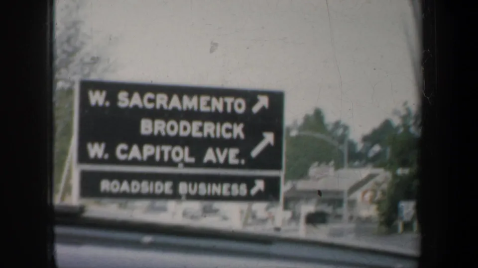 1950s California home-movie frame of a roadside directional sign viewed from inside a moving car, with window framing and soft film blur.
