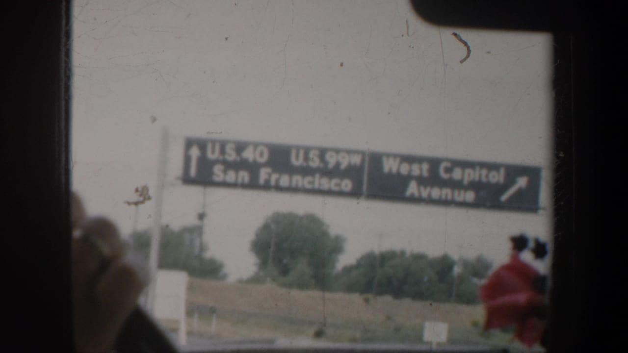 California road sign for U.S. 40 and U.S. 99W toward San Francisco, filmed through a vehicle window with a traveler partly visible at right.