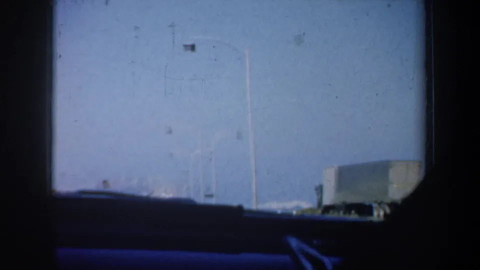 1957 San Francisco moving-car view of open sky, street light poles, and a pale roadside structure or vehicle in the distance.