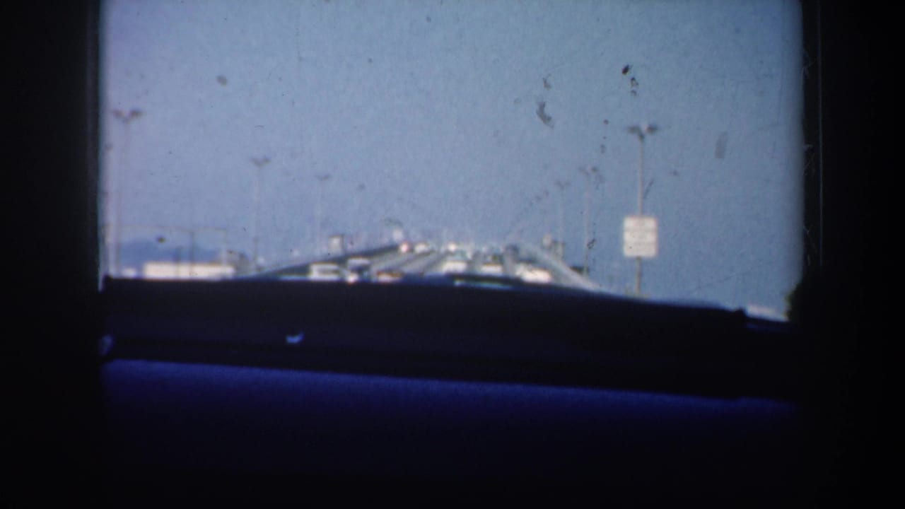 1957 San Francisco-area car-window view showing a dark interior, blue dashboard or seat edge, bridge rail, pale sky, and distant shoreline structures.